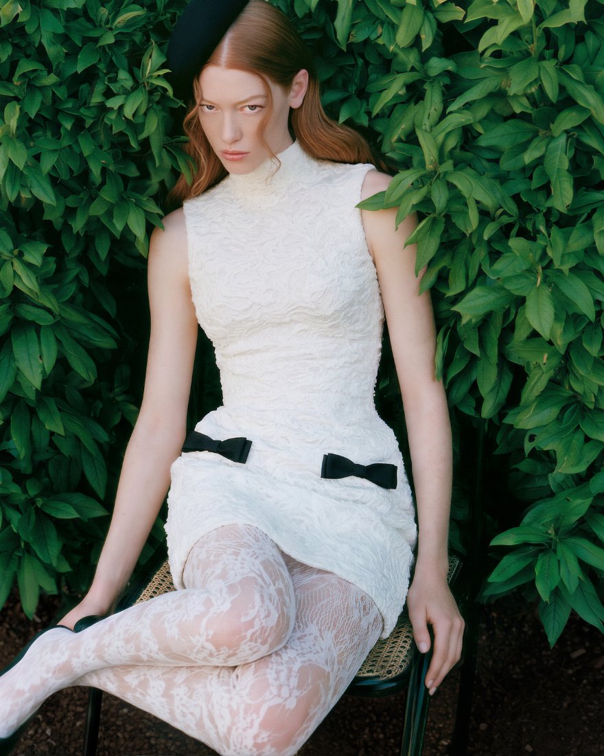 Model in a white mini dress with black bows and lace tights in front of a hedge