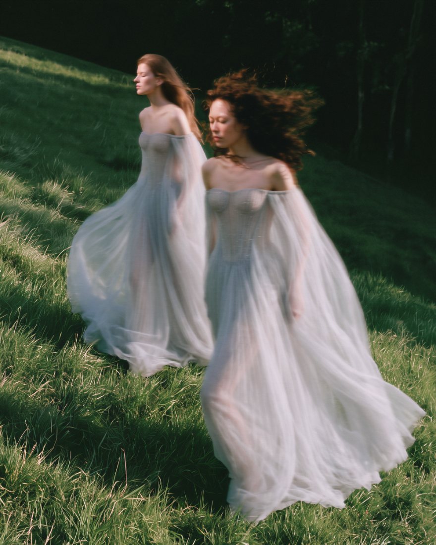 Two models in sheer off-shoulder gowns walking up a grassy hillside