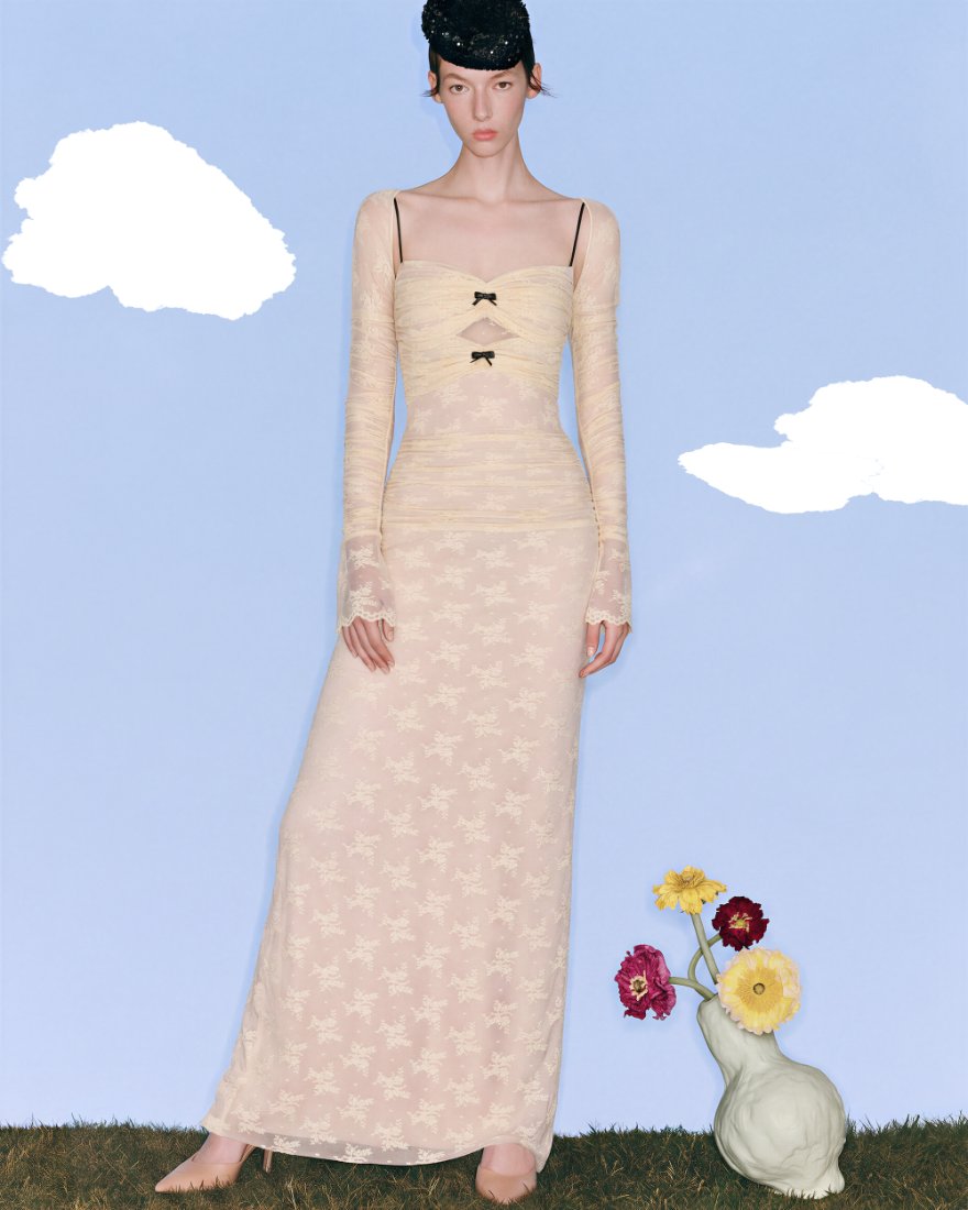 Model in a lace column dress against a painted blue sky with a vase of flowers